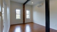 This single-floor space offers a compelling blend of industrial character and modern design. The interior showcases rich hardwood flooring that runs throughout, reflecting natural light and adding warmth to the overall aesthetic. A standout architectural feature is the exposed brick column that rises from floor to ceiling, providing a striking contrast against the clean white walls and serving as a reminder of the building's historic character. A bold dark structural beam spans the ceiling, adding to the industrial charm of the space. Natural light flows generously through multiple large, grid-paned windows, each framed in black — a detail that complements the space's contemporary edge while offering views of the surrounding greenery outside. Recessed lighting in the ceiling provides additional illumination, ensuring the space remains bright at any hour. A wall-mounted mini-split air conditioning unit ensures year-round climate comfort without compromising the clean lines of the interior. The high ceilings amplify the sense of openness and volume throughout. The combination of raw, textural elements like exposed brick and the dark beam alongside polished hardwood floors and bright white walls creates a balanced, loft-inspired ambiance that is both functional and visually engaging.