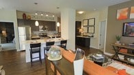 This single-floor residence offers a thoughtfully designed open-concept layout that seamlessly connects the kitchen, dining, and living areas into one cohesive living space. The modern kitchen is a standout feature, showcasing dark espresso cabinetry, stainless steel appliances, and a striking mosaic tile backsplash that adds texture and visual interest. A large kitchen island with light countertops provides ample prep space and doubles as a breakfast bar with stylish bar seating — ideal for casual dining and entertaining. Pendant lights hang above the island, complementing the track lighting overhead to create a warm, well-lit atmosphere. Adjacent to the kitchen, a dedicated bar or butler's area with matching dark cabinetry offers additional storage and serving functionality. The open living and dining areas are anchored by rich, dark hardwood flooring that runs throughout, adding warmth and sophistication to the space. Neutral walls provide a versatile backdrop, allowing the bold, contemporary furnishings — including an orange accent sofa — to stand out. Decorative wall art and carefully placed lighting enhance the overall ambiance, making the space feel curated and inviting. The layout is practical, stylish, and well-suited for modern everyday living.