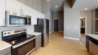This thoughtfully designed single-floor unit offers a contemporary living experience with a neutral color palette of warm grays and taupe tones throughout. The open-concept kitchen is a standout feature, boasting stainless steel appliances including a refrigerator, electric range, and microwave, complemented by a two-tone cabinet design with dark wood lower cabinets and crisp white uppers, topped with light countertops. Light hardwood-style flooring runs through the kitchen and entry areas, adding warmth and visual continuity. The generously sized living area is carpeted and filled with natural light, featuring a sliding glass door that opens to a private balcony with outdoor views. Two-panel white interior doors and clean white baseboards provide a polished contrast against the neutral walls. The bedroom is a comfortable, carpeted retreat with a ceiling fan, a bright window with blinds, and ample wall space. The bathroom is well-equipped with a full tub-shower combination, wood-look tile flooring, and classic white fixtures. The dollhouse view reveals a practical and efficient floor plan with a dedicated closet area and well-proportioned rooms, making the most of the available space throughout this move-in-ready unit.