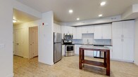This single-floor space offers a clean, contemporary design centered around a well-appointed kitchen. The kitchen is outfitted with a full suite of stainless steel appliances, including a refrigerator, range with oven, over-the-range microwave, and dishwasher. Crisp white shaker-style cabinetry lines the walls, providing ample storage, while a light gray subway tile backsplash adds a subtle texture and modern character. The countertops complement the overall neutral palette, and a single-basin sink is positioned beneath a window, enhancing the bright, airy feel of the space. Recessed lighting is installed throughout the ceiling, ensuring even, well-distributed illumination across the open layout. Light hardwood-style flooring flows seamlessly through the space, contributing to the clean and cohesive aesthetic. A wooden kitchen island or prep table adds a warm contrast to the otherwise light interior. The open layout connects the kitchen to the adjacent living area, creating a functional and inviting environment. Additional features visible include interior doors with a classic panel design, contributing to the overall polished and move-in-ready presentation of this thoughtfully designed space.