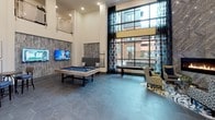 This expansive single-floor amenity lounge is defined by its soaring double-height ceilings, which lend an airy, grand atmosphere throughout the space. Large floor-to-ceiling windows dressed with bold patterned drapes flood the interior with natural light while offering views of the surrounding exterior. The flooring throughout is composed of oversized hexagonal tiles in a cool slate tone, providing a cohesive and modern foundation.

The centerpiece of the lounge is a dramatic floor-to-ceiling accent wall clad in textured chevron-patterned stone tile, housing a sleek linear gas fireplace. Surrounding the fireplace is a curated seating arrangement featuring patterned accent chairs and a glass coffee table atop a graphic area rug, creating an intimate gathering spot. Nearby, a secondary lounge nook offers a sofa, navy barrel chair, and abstract artwork displayed against the tall white walls.

A game area is anchored by a full-size billiards table alongside a mounted flat-screen television and wall-mounted cue rack. The reception area includes a curved front desk with decorative metallic wall art above and a mezzanine-level overlook with glass railings. Together, these zones create a sophisticated, well-appointed communal environment.