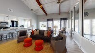 Located at 1800 William Kennerty Dr in Charleston, SC, this single-story community clubhouse offers a thoughtfully designed gathering space with a warm, inviting atmosphere. The main lounge area makes an immediate impression with its soaring vaulted ceiling accented by rich, exposed wood beams and white shiplap, lending a coastal-contemporary character to the space. Light wood flooring flows throughout, complementing the neutral gray walls and pops of bold orange and earthy gold in the furnishings. A central fireplace framed by a crisp white mantle serves as a focal point, paired with a mounted television and wall sconces for an elegant yet functional touch. Comfortable seating arrangements — including a dark sofa, upholstered armchairs, and round ottomans — encourage relaxed socializing, while a dedicated workstation provides a quiet spot for productivity. Large sliding glass doors and generous windows flood the space with natural light and offer views of the surrounding grounds. Adjacent to the lounge, a fitness center is visible behind full-height mirrored glass panels and is outfitted with exercise equipment, ceiling fans, and ample natural light. The kitchen features granite countertops, white cabinetry, stainless steel appliances, and a pass-through window that connects seamlessly to the main gathering area, making it well-suited for events and everyday use.