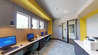 This business center is a thoughtfully designed, single-floor workspace that combines functionality with a vibrant, modern aesthetic. The space is immediately distinguished by its bold color palette — striking yellow accent walls and trim contrast against cool gray surfaces, creating an energetic yet focused atmosphere.

The main workstation area features a long, continuous white countertop running along one wall, accommodating multiple desktop computers paired with ergonomic teal rolling chairs. A row of sleek pendant lights suspended on red cords hangs above the workstation counter, providing focused task lighting. A series of windows along this wall invites natural light and offers views of the surrounding greenery, reducing the enclosed feel of the workspace.

A dedicated print and copy station occupies its own alcove, highlighted by a bold yellow rectangular frame accent on the wall. Dark cabinetry below the white countertop provides ample storage, while an HP all-in-one printer sits prominently for easy access.

The space also includes a glass-walled private conference or meeting room, offering a semi-enclosed area for more focused work or small group sessions. Patterned, tile-like carpeting in muted blue and gray tones grounds the space and ties together the overall design. The business center is a practical and visually engaging environment well-suited for productivity.