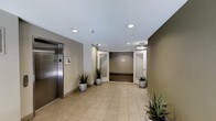 This lobby space presents a clean, professional atmosphere designed to provide a welcoming first impression. The flooring consists of large-format beige ceramic tiles that extend throughout the area, contributing to a bright and open feel. Recessed lighting is evenly distributed across the white ceiling, ensuring consistent, well-lit conditions throughout the space.

A stainless steel elevator with a modern control panel is prominently positioned along the left wall, providing convenient vertical access. The walls feature a neutral two-tone color scheme, combining warm beige tones on the lower sections with an olive green accent on the right, adding visual depth and contrast.

At the far end of the lobby, a curved architectural feature frames a set of glass-paneled doors, creating a natural focal point that draws the eye and guides movement through the space. This design element adds a subtle sophistication to the otherwise functional layout.

Potted snake plants are strategically placed throughout the corridor, introducing natural greenery that softens the commercial aesthetic. Overall, the space balances practicality with thoughtful design, creating an inviting and orderly environment suitable for a shared or commercial building entrance.