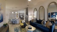 This well-appointed single-floor residence offers an open-concept layout that seamlessly connects the living, dining, and kitchen areas into one cohesive and functional space. The living area is anchored by a deep navy blue sofa accented with geometric patterned pillows, a bold yellow accent chair, and a faceted glass coffee table atop a richly colored area rug — creating a vibrant yet sophisticated aesthetic. A pair of tall arched mirrors adds depth and elegance to the living space.

The kitchen is outfitted with stainless steel appliances, including a refrigerator, range, and microwave, complemented by dark wood cabinetry and a light-toned countertop island with bar seating. Pendant lighting hangs above the island, providing both task lighting and a modern design touch. The open kitchen flows naturally into the dining and living zones, making it ideal for entertaining.

Throughout the space, light wood-style flooring contributes to a clean, contemporary look, while neutral white walls keep the interior bright and airy. High ceilings and recessed lighting enhance the sense of openness. The overall ambiance is modern and inviting, with a thoughtful blend of bold color accents and refined architectural details.
