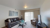This single-floor space centers around a well-appointed living room designed for comfort and functionality. The room features neutral-toned walls that create a clean, versatile backdrop, complemented by smooth white ceilings with a centrally mounted flush light fixture. A large sliding glass door dressed with vertical blinds serves as the room's focal point, allowing natural light to filter in while offering easy access to the exterior. The carpeted flooring adds warmth and softness underfoot throughout the living area. Seating is anchored by a dark leather sofa accented with a decorative pillow, paired with a tan upholstered armchair, forming a practical and inviting conversation area. A glass-topped coffee table with a sculptural metal base sits at the center, adding a contemporary touch. A small console table with a lamp provides additional ambient lighting, while a large abstract canvas in cool blue and grey tones adds a artistic accent to the wall. A flat-screen television is positioned to one side, completing the entertainment setup. The overall ambiance is calm and move-in ready, balancing modern accents with everyday livability.
