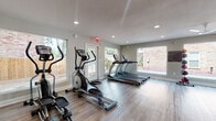 This fitness center is a thoughtfully designed single-floor exercise space that balances functionality with a clean, modern aesthetic. The room features wood-look flooring throughout, providing a durable and attractive surface underfoot, while neutral-toned walls and recessed lighting create a bright, energizing atmosphere.

The cardio section is well-stocked with multiple treadmills, elliptical cross-trainers, and a rowing machine, all positioned to take advantage of the abundant natural light streaming through the large windows that frame views of the surrounding outdoor environment. A wall-mounted flat-screen television provides entertainment during workouts.

The strength training area includes a multi-function cable and weight machine, a flat bench with a squat and pull-up rack, and a full dumbbell rack offering a range of free weights. A stability ball rounds out the available equipment, supporting a variety of exercise routines.

Glass panel dividers and a glass-framed entryway contribute to the open, airy feel of the space, while multiple exterior doors enhance ventilation and accessibility. The generous layout ensures ample room to move between equipment comfortably. Overall, the fitness center offers a comprehensive and inviting environment for both cardio and strength-focused workouts.