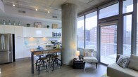 This single-floor urban loft showcases an industrial-modern aesthetic through a thoughtful combination of raw materials and contemporary finishes. A striking cylindrical concrete column anchors the open-plan layout, complementing the exposed concrete ceiling fitted with track lighting throughout. The kitchen features sleek white cabinetry, stainless steel appliances, and a rustic wooden dining table paired with industrial-style adjustable stools, blending utilitarian and modern design sensibilities seamlessly. Adjacent to the kitchen and dining area, a cozy sitting nook is furnished with a patterned accent chair and side table, positioned to take full advantage of the expansive floor-to-ceiling windows. These large windows flood the interior with abundant natural light while framing panoramic views of the surrounding urban skyline. Wide-plank hardwood flooring runs throughout, adding warmth that contrasts effectively with the industrial concrete elements above. The overall ambiance is open, airy, and urban — a well-balanced space that merges raw architectural character with modern comfort and functionality.