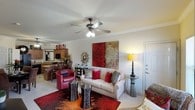 This single-story space offers an open-concept layout where the living area connects seamlessly to the kitchen, creating a cohesive and functional floor plan. The living room serves as the focal point, anchored by a neutral sofa adorned with decorative red and metallic pillows, complemented by a rich red accent chair that adds a bold pop of color. A striking large-scale artwork in warm red and gold tones dominates the main wall, reinforcing the space's vibrant color palette. A ceiling fan with an integrated light fixture is centrally mounted, providing both airflow and ambient lighting. Crown molding lines the perimeter of the ceiling, adding an architectural detail that lends a refined touch to the room. The flooring is topped with a vivid patterned area rug in red and earthy tones, grounding the seating arrangement. Decorative side tables, including a bold black-and-white striped accent table, add visual interest and contrast. The kitchen, visible in the background, features warm wood cabinetry, a breakfast bar with seating, and modern appliances, making it both practical and inviting. The overall ambiance is warm, energetic, and thoughtfully styled.
