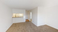 This single-floor unit offers a clean, functional layout designed for comfortable everyday living. The open-concept living area is finished with neutral wall-to-wall carpeting and smooth white walls, creating a bright and versatile space. A textured ceiling adds subtle character throughout the main living areas. Natural light filters generously through a large sliding glass door fitted with vertical blinds, illuminating the carpeted bedroom and providing a connection to the outdoors.

The kitchen is positioned adjacent to the living area and features a pass-through counter with a raised breakfast bar, white cabinetry extending to the ceiling for ample storage, a white range, and a sink. The kitchen floor transitions to hardwood-style flooring, offering a practical contrast to the carpeted living spaces. A short hallway connects the main living area to the bedroom and bathroom, maintaining a clear and organized flow throughout the unit.

The bathroom is outfitted with a full-size soaking tub and overhead shower with chrome fixtures, set against a clean white surround. The overall aesthetic of the space is neutral and move-in ready, with white walls and minimal detailing throughout that provide a versatile canvas for personalization.