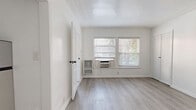This 210 square foot studio apartment in Los Angeles offers a well-designed, compact living space. The main living area features light gray wood-look flooring that extends throughout, creating a cohesive and spacious feel. Large windows with horizontal blinds allow ample natural light, brightening the space and providing a connection to the outdoors.

The kitchenette, while compact, is efficiently designed with white cabinetry, a stove, and necessary appliances. The bathroom, accessed through a white paneled door, boasts a clean, modern design with a pedestal sink, toilet, and what appears to be marble-look tile flooring.

The apartment's white walls and trim, including crown molding, contribute to its fresh and contemporary aesthetic. A wall-mounted air conditioning unit provides climate control. The space is ideal for a single occupant or couple seeking a minimalist, low-maintenance lifestyle in an urban setting. Despite its small footprint, the thoughtful layout and design elements maximize functionality and create a sense of openness in this cozy Los Angeles dwelling.