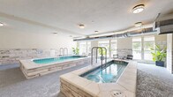 Located in Nashville, Tennessee, this indoor aquatic and wellness facility offers a well-appointed, single-floor space designed for relaxation and recreation. At its centerpiece is a generously sized indoor swimming pool with clear turquoise water, tiled in classic blue and surrounded by a smooth, light-toned deck. The pool reaches a depth of 3 feet 6 inches and is equipped with a safety railing for easy entry. Adjacent to the pool sits a raised hot tub, ideal for hydrotherapy and unwinding. A sauna room is also accessible, adding to the wellness-focused amenities of the space. The interior walls are clad in a whitewashed stacked-stone tile, lending a clean, spa-like aesthetic throughout. Lush tropical potted plants are strategically placed around the room, enhancing the serene atmosphere. A dedicated relaxation lounge area lines one side of the space, featuring cushioned lounge chairs positioned along a wall of large windows. These windows flood the area with natural light and offer views of the surrounding greenery and the Nashville skyline beyond. Exposed ductwork runs along the ceiling, adding an industrial contrast to the warm, resort-inspired design. Safety signage and a life ring are prominently displayed, ensuring a safe environment for all users.