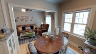 This charming single-floor residence exudes a cozy, welcoming ambiance rooted in classic architectural character. The open-concept layout seamlessly connects the dining area and living room, creating a fluid and functional living space. Crown molding and crisp white trim throughout add a timeless elegance to the warm, neutral-toned walls.

The dining area is anchored by a round hardwood table surrounded by white chairs and wicker seating, centered beneath natural light pouring in through large double-hung windows. A white radiator beneath the window adds to the home's traditional charm. The space is accented with decorative candle arrangements, lending a lived-in, inviting feel.

Flowing naturally into the living room, the space features rich hardwood flooring, a striped area rug, and comfortable seating including a brown leather sofa adorned with navy accent pillows and a coordinating recliner armchair. A green coffee table serves as a casual, eclectic focal point. A ceiling fan overhead and additional wall art and decorative accents complete the room's layered, homey aesthetic.

Together, these spaces offer a well-appointed, light-filled interior that balances comfort and classic design throughout.