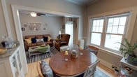This charming single-floor residence exudes a cozy, welcoming ambiance rooted in classic architectural character. The open-concept layout seamlessly connects the dining area and living room, creating a fluid and functional living space. Crown molding and crisp white trim throughout add a timeless elegance to the warm, neutral-toned walls.

The dining area is anchored by a round hardwood table surrounded by white chairs and wicker seating, centered beneath natural light pouring in through large double-hung windows. A white radiator beneath the window adds to the home's traditional charm. The space is accented with decorative candle arrangements, lending a lived-in, inviting feel.

Flowing naturally into the living room, the space features rich hardwood flooring, a striped area rug, and comfortable seating including a brown leather sofa adorned with navy accent pillows and a coordinating recliner armchair. A green coffee table serves as a casual, eclectic focal point. A ceiling fan overhead and additional wall art and decorative accents complete the room's layered, homey aesthetic.

Together, these spaces offer a well-appointed, light-filled interior that balances comfort and classic design throughout.