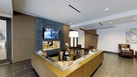 This single-floor space exudes a refined, contemporary aesthetic designed for both comfort and style. At the heart of the lounge area sits a striking floor-to-ceiling feature wall clad in textured dark stone tile, incorporating a sleek fireplace and a wall-mounted flat-screen television — a bold focal point that anchors the entire room. A generously sized sectional sofa with warm gold-toned wood paneling wraps around a set of brushed brass accent tables, creating an inviting and intimate seating arrangement. Soft accent pillows in muted tones add a layer of warmth to the neutral palette. Overhead, a large square pendant light with a white shade provides a soft, diffused glow, complemented by recessed ceiling lighting that brightens the space evenly. Large-format tile flooring in a cool gray tone grounds the room with a clean, polished finish. A distinctive angled wood architectural element frames the transition to an adjacent area, adding visual interest and a touch of organic warmth to the otherwise modern interior. Additional seating with upholstered chairs is visible beyond, suggesting a thoughtfully zoned layout that encourages both social interaction and quiet retreat. The overall ambiance is sophisticated, welcoming, and meticulously curated.