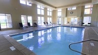 This indoor aquatic facility is housed within a single-story structure characterized by soaring ceilings and an abundance of natural light. Large windows line the walls on multiple sides, flooding the interior with daylight and offering views of the surrounding landscape, creating an open and airy atmosphere despite being fully enclosed.

The centerpiece of the space is a generously sized rectangular swimming pool with crystal-clear blue water, framed by a clean tiled deck. Handrails at the pool's edge provide convenient entry and exit points. Adjacent to the main pool is a hot tub, offering a relaxing alternative for guests.

Along the poolside deck, a row of black sling-back lounge chairs and small side tables provides a comfortable area for relaxation and socializing. The neutral-toned walls and light-colored flooring complement the bright, welcoming environment.

The high clerestory windows not only maximize natural illumination throughout the day but also contribute to the facility's spacious, resort-like character. Overall, the space presents a clean, well-appointed aquatic environment suitable for both recreational swimming and leisurely relaxation.