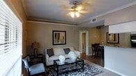 This single-floor space offers a thoughtfully designed interior that balances comfort and style. The living area serves as the heart of the home, featuring warm neutral-toned walls accented by crisp white crown molding that adds a refined architectural touch. A decorative ceiling fan with light fixture provides both ambiance and functionality, while HVAC vents ensure year-round comfort.

The living room is furnished with a plush light-colored sofa adorned with coordinating throw pillows, complemented by a sleek glass-top coffee table resting on a bold, patterned area rug in dark brown and white tones. Decorative spherical accents on the coffee table add an artistic element, while framed wall art and a classic table lamp complete the sophisticated aesthetic.

Adjacent to the living area, a glimpse into the dining space reveals dark wood bar-height seating and a counter area, suggesting a seamless flow between living and entertaining spaces. The hardwood-style flooring in the dining area contrasts beautifully with the carpeted living room, creating distinct yet connected zones. A hallway visible in the background hints at additional private spaces beyond, making this a well-organized and inviting single-floor residence.