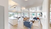 Located at 1510 Valentia Street in Denver's 80220 neighborhood, this community room is a well-appointed, single-floor gathering space designed to serve a variety of communal needs. The open-plan layout is anchored by light wood-tone flooring and a crisp white ceiling with recessed tray details and modern pendant and flush-mount lighting throughout.

A fully equipped kitchen lines one wall, featuring an expansive dark quartz island with bar seating for multiple guests, white shaker-style upper and lower cabinetry, a subtle subway tile backsplash, stainless steel appliances including an oven and microwave, and a deep utility sink — making the space well-suited for events and gatherings.

The lounge area is furnished with a leather sofa, blue upholstered accent chairs, and a coffee table arranged on a neutral area rug, complemented by a wall-mounted flat-screen television and built-in shelving with storage cabinetry. A dedicated dining zone offers white tables and modern wood chairs, ideal for meetings or casual dining.

Floor-to-ceiling windows spanning multiple walls flood the interior with abundant natural light and provide open views of the surrounding Denver streetscape and tree-lined neighborhood. The overall ambiance is clean, modern, and welcoming — a functional yet comfortable community hub.