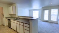 This single-floor residence offers a clean and functional open-concept layout that seamlessly connects the kitchen and living areas. The kitchen features white cabinetry with multiple drawers and cabinet doors providing generous storage, complemented by dark speckled countertops that extend along a peninsula-style breakfast bar. A stainless steel sink is integrated into the counter, adding a practical touch to the space. The peninsula serves as a natural dividing point between the kitchen and the adjoining living area, creating a sense of flow while maintaining distinct zones for cooking and relaxing. The living area is finished with plush wall-to-wall carpeting that adds warmth and comfort underfoot. Large windows on two walls flood the space with abundant natural light, creating a bright and airy ambiance throughout. The neutral white walls and light-toned ceilings further enhance the open, spacious feel of the interior. The overall design is straightforward and practical, making it well-suited for everyday living while offering a blank canvas for personalization and décor.