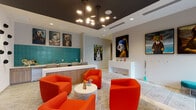 Located at NE Sixth Street in Fort Lauderdale, this single-floor pet amenity space combines functional design with a lively, gallery-like aesthetic. The layout is divided into two distinct zones: a social lounge area and a dedicated pet wash and grooming room, connected by a corridor with interior windows that offer views between the two spaces.

The lounge area makes an immediate visual impact with bold red barrel chairs arranged around a round marble-top coffee table, set atop striking black-and-white geometric patterned tile flooring. A dark-painted ceiling tray overhead is fitted with a sculptural multi-arm chandelier, creating contrast against the bright white walls. A wall-mounted flat-screen television sits above warm wood cabinetry, and a service counter with teal subway tile backsplash runs along one side. Throughout the lounge, large-format pop-art-style dog portraits line the walls, adding a whimsical, curated character to the space.

The pet wash room features floor-to-ceiling white subway tile, a raised wash basin, and decorative patterned wallcovering near the ceiling. Stainless steel sinks, wood cabinetry, and built-in counters provide practical grooming functionality. Interior black-framed windows connect this area to the main lounge, maintaining an open and cohesive feel across the entire space.