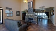 This single-floor space makes a striking first impression with its soaring ceilings and open-concept design that encourages a natural flow between living and dining areas. The centerpiece of the living room is a grand stacked-stone fireplace, rising dramatically toward the ceiling and anchored by substantial stone columns on either side. A framed mirror mounted above the fireplace mantel adds depth and elegance to the composition. Rich, large-format tile flooring in warm earth tones grounds the space and complements the neutral, sun-warmed wall tones throughout. The living area is furnished with dark leather seating arranged around a classic wooden coffee table, creating a comfortable and inviting gathering space. A decorative ceiling light fixture adds a warm glow overhead. Adjacent to the living area, the dining space benefits from ample natural light streaming through expansive glass doors or windows, offering views to an outdoor area. A ceiling fan serves this secondary zone, enhancing comfort. Decorative framed artwork and a dark wood armoire add character and a sense of curated style. The overall ambiance is warm, substantial, and refined, with a Southwestern-inspired aesthetic running throughout.