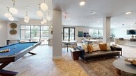 Located at 301 Silveron in Flower Mound, Texas, this thoughtfully designed single-floor amenity space offers a generous and versatile layout ideal for socializing and relaxation. The open-concept design seamlessly connects a fully equipped kitchen and lounge area with multiple gathering zones, all unified by striking geometric-patterned hardwood flooring throughout.

The kitchen area serves as a centerpiece, featuring a large white island with leather-and-brass bar seating, sleek gray cabinetry, a stainless steel refrigerator, and a stunning branch-inspired chandelier overhead. A textured mosaic tile accent wall adds visual depth and sophistication to the space.

The expansive living area is divided into distinct zones: a cozy lounge with a wall-mounted TV framed by a warm, textured wallcovering; a game room with a full-size billiards table illuminated by a cluster of globe pendant lights in brass fixtures; and casual dining areas with round tables and modern chairs. A dedicated coffee and refreshment bar with counter seating runs along a window wall, bathed in natural light.

Large windows and glass-paneled doors throughout the space invite abundant daylight and offer views of the outdoor surroundings. The overall ambiance is modern and inviting, with a cohesive palette of warm neutrals, soft blues, and gold accents that lend an upscale, welcoming character to every corner.