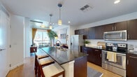 Located in the heart of downtown Orlando, this 1-bedroom, 1-bathroom residence offers a well-designed open-concept layout that seamlessly connects the kitchen, dining, and living areas. The kitchen is a standout feature, anchored by a large granite-topped island with bar seating, dark espresso cabinetry, a white subway tile backsplash, stainless steel appliances, and pendant lighting overhead. Light hardwood floors flow throughout the main living areas, complementing the exposed concrete ceiling that lends an urban, industrial character to the space. The living and dining area is bathed in natural light through expansive floor-to-ceiling windows that frame sweeping views of the Orlando skyline. The bedroom continues the city view theme with generous glazing, carpet flooring, and ample closet storage. The bathroom is finished with a dark vanity topped in granite, white tile walls, and a tub-shower combination. A dedicated hallway leads to additional closets, a laundry area, and a utility room, providing practical storage solutions. Three closets throughout the unit ensure organizational flexibility. The overall ambiance is modern and urban, with a clean, contemporary palette that highlights the architectural details and cityscape views.