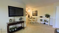 This single-floor home offers a comfortable and cohesive living environment designed for everyday ease. The open-concept layout connects the living area seamlessly to the dining space, creating a sense of openness and flow throughout the main living zone. The living area features wall-mounted flat-screen television paired with a dark wood console table, complemented by decorative greenery that adds a touch of warmth and character. Soft, neutral-toned carpeting runs throughout, contributing to the cozy and inviting ambiance of the space. The dining area is anchored by a round table with white Windsor-style chairs, lending a casual yet charming aesthetic. A ceiling fan with integrated lighting hangs above, providing both air circulation and ambient illumination. The walls are finished in a soft, warm neutral tone that enhances the brightness of the interior. A decorative framed artwork and a circular mirror accent the dining space, adding visual interest to the walls. A light wood console table along the dining perimeter provides additional storage and display space. Overall, the home presents a tidy, well-appointed interior that balances functional living with comfortable, homey styling.
