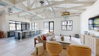 This single-story space showcases a coastal-inspired design defined by soaring vaulted ceilings with exposed white-painted wooden beams and visible ductwork, lending an airy, loft-like character to the interior. Large black-framed windows and glass doors flood the space with abundant natural light while offering views of the surrounding outdoor greenery.

The open-plan layout connects several distinct functional zones. The main lounge area features generous seating, including a substantial wood-framed sofa, rounded wicker accent chairs, and layered jute rugs atop large-format light tile flooring. A wall-mounted flat-screen television sits atop a whitewashed cabinet, complemented by a bold turquoise and yellow art piece. A beaded chandelier adds an elegant, coastal focal point overhead.

Adjacent to the lounge is a stylish bar area, distinguished by blue cabinetry, patterned tile flooring, and counter-height wicker barstools. A separate dining nook near a glass entry door provides a round white pedestal table with rattan director's chairs and framed artwork above.

Throughout, shiplap wall paneling reinforces the coastal aesthetic, while black-framed glass interior doors add a contemporary contrast. The space presents a welcoming, resort-inspired ambiance ideal for social gatherings.
