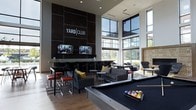 The Yard Club at 800 Baldwin Dr in Grandview Heights, Ohio is a thoughtfully designed communal social space that blends contemporary style with an inviting, energetic atmosphere. The single-floor layout is anchored by soaring ceilings and expansive floor-to-ceiling windows that flood the space with natural light and offer open views of the surrounding exterior, creating an airy, spacious feel.

At the heart of the space is a sleek bar area, finished in dark, large-format tile cladding that extends dramatically up the wall as a bold focal feature. Two wall-mounted flat-screen televisions flank the Yard Club signage above the bar, making it an ideal spot for watching sports or events. Bar-height seating with red-cushioned stools lines the counter, providing a casual perch for socializing.

Adjacent to the bar, plush lounge seating — including dark gray sofas and accent chairs with colorful throw pillows — is arranged around low side tables, encouraging relaxed conversation. Warm hardwood-style flooring runs throughout, complementing the modern, dark-toned design palette.

A full-size pool table serves as a central recreational anchor, inviting friendly competition. The overall ambiance is sophisticated yet approachable, combining upscale finishes with a welcoming, social energy that makes the Yard Club a standout amenity space.