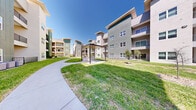 This contemporary residential community is defined by its thoughtfully designed outdoor common areas set between multiple three-story buildings. The architecture features a clean, modern aesthetic with a neutral stucco and panel exterior accented by teal and green facade details, large windows, and private balconies that overlook the shared grounds below.

The central courtyard serves as the heart of the community, offering a generous expanse of well-maintained lawn that connects the surrounding buildings. A covered outdoor pavilion with a wood-paneled ceiling, ceiling fan, and built-in grill station provides a dedicated space for outdoor entertaining and social gatherings. Smooth concrete walkways wind through the landscaped grounds, guiding residents between buildings and communal areas with ease.

Newly planted trees and manicured shrubs are distributed throughout the green space, adding natural texture and shade as the landscaping matures. Additional outdoor seating areas with fenced patios are accessible at ground level, offering private outdoor space for ground-floor units. The community also features dedicated HVAC infrastructure positioned along the building exteriors. Under clear, open skies, the overall ambiance of the outdoor space is bright, open, and inviting, reflecting a well-planned community environment designed for both relaxation and everyday living.