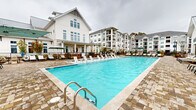 Located at 11023 Moonbug Ct in Charlotte, NC, this single-floor community amenity space offers a generously sized clubhouse and resort-style pool area with a distinctly coastal aesthetic. The clubhouse is defined by its dramatic vaulted ceilings with exposed white-painted beams, flooded with natural light through expansive clerestory windows fitted with plantation shutters. Elegant cage-style pendant lanterns hang throughout, complementing the light stone tile flooring and bright, white shiplap wall paneling.

The main gathering area features spacious sectional seating arrangements in neutral tones, centered around a freestanding double-sided fireplace, creating an inviting social hub. A separate seating nook includes a kitchenette with stainless steel appliances, ideal for entertaining. Wicker dining sets positioned near the windowed walls offer additional casual seating with views of the outdoor area.

The outdoor pool deck is a standout feature, showcasing a large rectangular swimming pool with striking blue water, surrounded by an expansive cobblestone patio lined with rows of cushioned lounge chairs and shaded seating areas. Manicured evergreen shrubs and landscaped planters add a refined touch to the outdoor environment, which looks out toward the surrounding multi-story residential buildings.