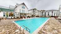 Located at 11023 Moonbug Ct in Charlotte, NC, this single-floor community amenity space offers a generously sized clubhouse and resort-style pool area with a distinctly coastal aesthetic. The clubhouse is defined by its dramatic vaulted ceilings with exposed white-painted beams, flooded with natural light through expansive clerestory windows fitted with plantation shutters. Elegant cage-style pendant lanterns hang throughout, complementing the light stone tile flooring and bright, white shiplap wall paneling.

The main gathering area features spacious sectional seating arrangements in neutral tones, centered around a freestanding double-sided fireplace, creating an inviting social hub. A separate seating nook includes a kitchenette with stainless steel appliances, ideal for entertaining. Wicker dining sets positioned near the windowed walls offer additional casual seating with views of the outdoor area.

The outdoor pool deck is a standout feature, showcasing a large rectangular swimming pool with striking blue water, surrounded by an expansive cobblestone patio lined with rows of cushioned lounge chairs and shaded seating areas. Manicured evergreen shrubs and landscaped planters add a refined touch to the outdoor environment, which looks out toward the surrounding multi-story residential buildings.
