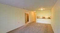 This single-floor space features an open-concept layout that connects the living area to the kitchen. The living room is finished with wall-to-wall beige carpeting, creating a warm and neutral foundation throughout. Smooth, light-colored walls keep the interior feeling clean and bright, complemented by flush-mount ceiling light fixtures that provide consistent overhead illumination.

A pass-through counter separates the kitchen from the main living area, offering a functional and connected layout. The kitchen is equipped with white appliances, including a refrigerator and range, and features a dark countertop along the pass-through. The design promotes ease of interaction between the cooking and living spaces.

A doorway visible from the main living area leads to additional rooms, suggesting a straightforward and practical floor plan. The transition areas are finished with consistent flooring and neutral-toned walls, maintaining a cohesive aesthetic throughout the space. Overall, the interior presents a simple, functional design with a neutral color palette suited for a variety of furnishing styles.
