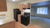 This single-floor residence offers a practical and functional layout designed for comfortable everyday living. The kitchen serves as a central feature of the space, outfitted with warm-toned wood cabinetry providing ample storage through both upper and lower cabinets. Black appliances, including a refrigerator, electric range with overhead ventilation, and a double-basin sink set into a granite-style countertop, give the kitchen a cohesive, modern look. The kitchen is finished with hardwood-style flooring that adds warmth and is easy to maintain. The kitchen opens directly into the adjoining living area, which transitions to plush carpeting, creating a comfortable and welcoming environment. A doorway from the kitchen provides a clear sightline into the bedroom, which also features carpeted flooring and benefits from natural light through a window. The living area similarly receives natural light, contributing to an airy and bright ambiance throughout the unit. Neutral wall tones in soft beige run consistently through the space, lending a clean and versatile backdrop. Practical details such as floor vents, standard white doors, and well-placed lighting fixtures complete the interior, making this a straightforward and move-in-ready space.