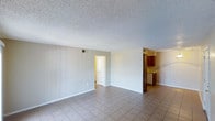 This single-floor space offers a straightforward and functional layout designed for comfortable everyday living. The main living area is generously proportioned, featuring neutral grey walls, light ceramic tile flooring laid in a classic grid pattern, and a textured popcorn ceiling that adds subtle dimension overhead. The open feel of the room is enhanced by clean sightlines and minimal visual clutter, making the space feel bright and airy.

Toward the rear, the living area flows naturally into a connected kitchen space. The kitchen is appointed with warm, rich-toned wood cabinetry and includes a full-size refrigerator, providing ample storage and practical functionality. A countertop workspace is visible, offering room for meal preparation. The kitchen area is distinguished by slightly warmer lighting compared to the main living space, creating a welcoming contrast.

A doorway visible from the main living area leads further into the unit, suggesting additional rooms beyond — likely a bedroom or bathroom — offering privacy and separation from the common living spaces. HVAC vents are integrated into both the walls and ceiling, ensuring climate control throughout. White baseboards trim the perimeter of the rooms, providing a clean, finished look. Overall, the space presents a practical and well-maintained interior ready for personalization.