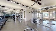Located at 401 S Main St in Akron, Ohio, this well-equipped fitness center offers a single-floor layout designed to accommodate a full range of workout needs. The space features an open, airy design with polished concrete flooring throughout, lending the facility a clean and modern aesthetic. Multiple ceiling fans are mounted overhead alongside industrial-style track lighting and exposed ductwork, contributing to a functional yet contemporary atmosphere.

The strength training area is outfitted with a variety of cable machines and multi-station cable systems, free-weight benches, and an extensive dumbbell rack spanning a full weight range. Full-length mirrored walls line portions of the space, enhancing the sense of openness and allowing users to monitor their form during exercise. A dedicated cardio zone houses a row of treadmills and elliptical machines positioned along large windows, which provide ample natural light and views of the outdoor surroundings.

A wall-mounted television is positioned above the free-weight area for entertainment during workouts. A small kitchenette or storage cabinet is visible along one wall, adding convenience to the space. Large floor-to-ceiling windows and a glass door along one side of the facility open to an outdoor patio area, further brightening the interior and providing a seamless connection to the exterior environment.