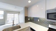 This single-floor space showcases a thoughtfully designed kitchen that serves as a central feature. The kitchen is outfitted with high-gloss white upper cabinetry extending toward the ceiling, maximizing storage while contributing to a clean, contemporary aesthetic. The cabinetry is complemented by crisp white lower cabinets with simple bar-pull hardware, maintaining a cohesive and minimalist look throughout.

A standout design element is the dark olive-green subway tile backsplash, which runs the full length of the counter walls, adding texture and contrast against the bright white surfaces. The countertops are light-colored and provide generous workspace, wrapping around in an L-shaped configuration. A stainless steel undermount sink with a modern faucet is positioned to one side, and a built-in dishwasher is integrated beneath the counter.

Recessed ceiling lighting provides focused illumination throughout the kitchen. The space opens into an adjacent living area, which benefits from natural light streaming through a sliding glass door or large window fitted with white roller blinds. The overall ambiance is bright, clean, and modern, with a neutral palette accented by the bold backsplash tile.