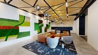 This contemporary lobby is a thoughtfully designed communal space that balances bold artistic expression with functional amenities. The defining feature of the room is an expansive moss wall installation spanning a large portion of one wall, composed of varied textures and vibrant shades of green, orange, and white arranged in an eye-catching, modular pattern that brings a natural, biophilic element to the modern interior.

The ceiling is an architectural highlight in itself, featuring a geometric grid of warm wood panels framed by black structural beams, with integrated linear lighting and a cascading cluster of mirrored chrome globe pendant lights that serve as a dramatic focal point. The open floor plan allows the lounge area to breathe, anchored by a substantial tan leather sectional sofa, rounded upholstered accent chairs in soft grey, and a patterned area rug in deep navy and black tones that grounds the space.

Polished concrete floors extend throughout, lending an industrial edge that complements the refined furnishings. Near the entrance, a dedicated mail and package center with black multi-unit mailboxes is neatly integrated into the layout. Elevator access and a well-lit entry corridor provide seamless connectivity. Large windows allow natural light to filter in, enhancing the overall sense of openness and contributing to the lobby's welcoming, design-forward ambiance.