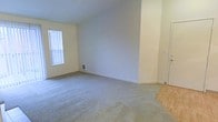 This single-floor residential space offers a clean, functional layout designed for comfortable everyday living. The main living area is finished with wall-to-wall carpet in a neutral tone, providing a soft and practical underfoot surface that contributes to the room's understated, move-in-ready aesthetic. The walls are painted in a light, warm off-white, creating a bright and open atmosphere throughout.

A prominent window fitted with vertical blinds allows natural light to filter into the space, illuminating the room and offering flexibility in light control. A floor-level ventilation register is positioned along the baseboard, indicating a ducted climate control system. The transition from carpet to a hardwood or laminate flooring surface is visible at the room's edge, hinting at a multi-surface floor plan that may include a kitchen, hallway, or entry area just beyond the main living space.

Clean white baseboards frame the room and complement the overall neutral palette. A light switch and utility features are neatly integrated into the walls, maintaining a streamlined appearance. The space presents a simple, adaptable interior that serves as a versatile foundation for a range of furnishing and decorating styles.