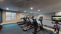 This fitness center is thoughtfully designed to provide a comfortable and fully functional workout environment. The space is anchored by a diverse array of cardio equipment, including treadmills, elliptical trainers, a stationary upright bike, a spin bike with an integrated display screen, and a rowing machine — offering users a comprehensive range of exercise options. A large wall-mounted mirror spans one side of the room, enhancing the sense of openness and allowing users to monitor their form during workouts. The flooring features a durable, patterned teal-blue carpet that adds a modern aesthetic to the space. Recessed lighting combined with multiple ceiling fans ensures the room remains well-lit and well-ventilated throughout use. Natural light filters in through several windows, which offer views of the surrounding building exterior. The walls are painted in a clean white on one side and a deep navy-teal on the accent wall, creating a dynamic yet focused atmosphere. A glass-paneled door provides access to the adjacent corridor, and occupancy signage is posted near the entrance. The overall design is clean, functional, and purposeful, making this fitness center a practical amenity for daily use.