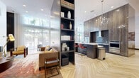 Located at 98 N 1st St in downtown San Jose, California, this stylish single-floor communal space offers a generous, open-plan layout combining a fully equipped kitchen and a spacious living and lounge area. Soaring ceilings throughout amplify the sense of openness, while light-toned herringbone hardwood flooring adds warmth and visual continuity across the entire floor.

The kitchen is a standout feature, anchored by a large central island with a white quartz countertop and integrated sink. Dark wood cabinetry lines the perimeter, complemented by stainless steel appliances including a full-size refrigerator, range, and oven. An elegant multi-arm chandelier overhead adds a refined touch. A vibrant mosaic tile accent wall in the dining area adds color and texture, serving as a bold backdrop for casual seating arrangements.

The living area is thoughtfully curated with mid-century modern furnishings, including structured wood-framed armchairs, a plush sofa, and layered area rugs in warm, earthy tones. A wall-mounted flat-screen TV and tall built-in shelving provide both function and style. A pool table positioned near floor-to-ceiling windows invites recreation, while sheer drapery filters abundant natural light and frames views of the surrounding outdoor greenery.