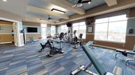 This fitness center offers a thoughtfully designed workout environment that balances functionality with comfort. The space is finished with a warm, two-tone color scheme of tan and soft blue-gray on the walls, complemented by a tray ceiling detail that adds architectural interest and a sense of height to the room. Ceiling fans are strategically positioned throughout to ensure consistent air circulation during workouts.

Large windows line the walls, flooding the space with abundant natural light and providing open views of the surrounding exterior, creating an airy and motivating atmosphere. The flooring features a multicolored striped carpet in shades of blue, teal, and gold, adding a dynamic visual element to the space.

The fitness center is well-stocked with cardio equipment, including stationary bikes, elliptical machines, and additional exercise equipment arranged to allow ample space for movement between machines. Multiple wall-mounted flat-screen televisions are positioned at comfortable viewing angles throughout the room, enhancing the workout experience. The overall ambiance is clean, bright, and inviting, making it a practical and pleasant space for daily exercise.
