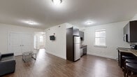 This thoughtfully designed 3-bedroom, 3-bathroom unit offers a functional and modern living experience across a single floor. The open-concept kitchen and living area serves as the heart of the home, featuring rich hardwood-style flooring, crisp white walls, and ample natural light from multiple windows. The kitchen is equipped with a full suite of stainless steel appliances — including a refrigerator, electric range, microwave, and dishwasher — complemented by dark espresso cabinetry, granite-style countertops, a tile backsplash, and a built-in breakfast bar with seating. The living area provides a comfortable, open layout with wall-mounted TV capability. Each of the three bedrooms is carpeted, offering a warm and quiet retreat, with direct access to their own bathrooms — making each room an en-suite. The bathrooms are finished with neutral large-format tile, glass-enclosed walk-in showers, wall-mounted vanities with modern fixtures, and framed mirrors with overhead lighting. The overall aesthetic is clean and contemporary, with a neutral color palette throughout that creates a cohesive and inviting atmosphere well-suited for shared or individual living.
