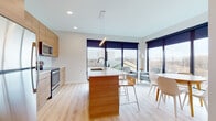 This thoughtfully designed single-floor residence showcases a clean, contemporary aesthetic centered around functionality and modern living. The open-concept main area seamlessly integrates the kitchen, dining, and living spaces beneath recessed lighting and a sleek pendant fixture. The kitchen is equipped with stainless steel appliances — including an electric range and built-in microwave — flat-front light wood cabinetry, and a white quartz countertop that extends to a center island with bar seating. Large floor-to-ceiling windows flood the space with abundant natural light and provide expansive outdoor views, while light hardwood-style flooring runs throughout. The bedroom features a standout built-in Murphy bed system framed by warm wood slat paneling and a matching slatted ceiling accent, offering a seamlessly convertible space that shifts between a comfortable sleeping area and a compact lounge with a built-in sofa and side tables. The bathroom is well-appointed with a warm wood vanity, white quartz countertop, a framed mirror, and a glass-enclosed walk-in shower. A dedicated laundry area houses a full-size stacked washer and dryer unit for added convenience. The overall ambiance is bright, warm, and efficiently designed.