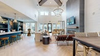 This thoughtfully designed single-floor communal space blends contemporary and transitional design elements across a generously sized open layout. The most striking architectural feature is the soaring vaulted ceiling, accented by exposed dark wood beams and clerestory windows that flood the interior with abundant natural light. A large tiered chandelier anchors the main lounge area, adding an elegant focal point overhead.

The primary living zone centers around a floor-to-ceiling tile-clad fireplace wall, complete with a linear hearth and wall-mounted television, surrounded by an eclectic mix of upholstered sofas, leather chairs, and woven accent seating arranged on a patterned area rug. Light-toned herringbone-patterned tile flooring extends throughout, unifying the space visually.

A dedicated kitchenette area features a large granite-topped island with bar seating, teal cabinetry, open shelving, a beverage refrigerator, exposed brick backsplash, and pendant lighting—making it ideal for gatherings. Adjacent dining and casual seating areas offer additional flexibility, including a round dining table near windows overlooking an outdoor area. Sliding barn doors, dark-framed glass entry doors, and teal accent walls complete the refined yet welcoming aesthetic.
