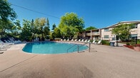 This outdoor pool facility in New Berlin, Wisconsin offers a well-appointed recreational space designed for relaxation and leisure. At the heart of the area is a generously sized oval swimming pool with clear, blue water, accessible via stainless steel entry rails and marked with depth indicators for safety. The pool is surrounded by a broad concrete sundeck that provides ample space for movement and socializing. Lining the perimeter of the deck is an extensive collection of white lounge chairs, offering plenty of seating for sunbathing. Shaded dining areas with umbrella-covered tables and chairs are positioned throughout the deck, providing comfortable spots for refreshments and conversation. The pool area is fully enclosed by a chain-link fence with a gated entry, ensuring security and privacy. Mature deciduous and evergreen trees frame the entire space, creating a natural, park-like atmosphere that offers both shade and visual appeal. Colorful landscaping with flower beds and mulched borders adds to the attractive setting. The backdrop features a multi-story brick residential building with balconies, reflecting the community-oriented nature of this amenity. Under open skies, this pool area presents an inviting and functional outdoor retreat.