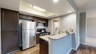 Located in West Valley City, Utah, this efficiently designed single-floor unit makes excellent use of its compact footprint. The kitchen serves as a central highlight, featuring dark espresso cabinetry, stainless steel appliances including a full-size refrigerator and range, and a peninsula with a built-in sink and light countertops. A recessed ceiling detail above the kitchen adds architectural interest and enhances the sense of openness. Durable wood-look flooring runs throughout the main living areas, providing a cohesive and easy-to-maintain surface. The two living areas offer flexible use of space, with one featuring sliding glass doors that open to a private balcony, allowing natural light to flow in and providing outdoor views. The bathroom is finished with wood-look flooring, neutral gray walls, and a vanity with dark cabinetry, maintaining a consistent design aesthetic. A dedicated laundry area houses a stacked washer and dryer unit, adding everyday convenience. A closet provides additional storage. The unit's neutral color palette of soft grays and warm wood tones throughout creates a calm, cohesive ambiance well-suited for comfortable everyday living.