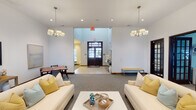 Located at 7247 Vista Dr in Indianapolis, Indiana, this single-floor community clubhouse offers a thoughtfully designed gathering space that balances comfort with style. The main lounge area is anchored by a dramatic floor-to-ceiling stone fireplace, complete with a rustic wooden mantle and hearth, serving as the room's defining architectural centerpiece. Soaring ceilings amplify the sense of openness, while multiple pendant chandeliers with drum shades cast warm, even lighting throughout. The seating area is furnished with cream-toned sofas and soft blue accent chairs arranged around a central coffee table, creating an inviting atmosphere for socializing or relaxing. A secondary lounge nook features a wall-mounted flat-screen television, additional seating, and its own chandelier, providing a more intimate setting within the space. Dark-framed French doors appear throughout, offering views to the outdoors and allowing natural light to filter in. A casual dining area with a wooden table and chairs adds functional versatility near the entryway. Neutral carpeting and crisp white walls provide a clean, cohesive backdrop that ties the various areas together. The overall ambiance is warm, polished, and welcoming — well-suited for community gatherings and everyday use alike.