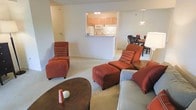This single-floor residence is designed with an open-concept layout that seamlessly connects the living, dining, and kitchen areas. The living room is a welcoming space, featuring plush seating and neutral-toned carpeting that adds warmth underfoot. A central coffee table anchors the seating arrangement, creating an inviting area for relaxation and conversation. Adjacent to the living area, the dining space accommodates a full table and chairs, making it ideal for everyday meals or entertaining. The kitchen is defined by a raised pass-through counter that opens to the main living area, enhancing the sense of connectivity throughout the space. Light wood cabinetry lines the kitchen walls, offering ample storage, while under-cabinet lighting and a pendant light above the pass-through add a practical yet decorative touch. The neutral color palette carried throughout the space — from the soft wall tones to the carpeted floors — creates a cohesive and calming ambiance. The open flow between rooms makes the overall layout feel spacious and well-proportioned, well-suited for comfortable day-to-day living.
